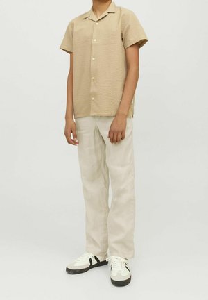 Chemise beige à manches courtes avec une finition texturée, boutonnée sur le devant, associée à un pantalon clair et des baskets noir et blanc.