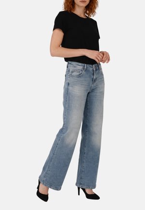 Vrouw draagt lichtblauwe jeans met wijde pijpen, zwart kort mouwtopje en zwarte hoge hakken, staand tegen een effen achtergrond.