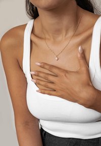Złocony naszyjnik z perłowym wisiorkiem, z delikatnym łańcuszkiem. Modelka ma na sobie białą dopasowaną bluzkę na ramiączkach i ma neutralny lakier do paznokci.