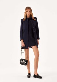 Cappotto di lana blu navy con colletto alto, bottoni frontali e tasche, indossato sopra un abito aderente. Borsa trapuntata nera e scarpe nere lucide.