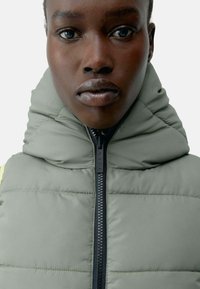 Lys grå puffer vest med høy krage og svart glidelås. Stoffet har en glatt tekstur og kviltet design med en slank silhuett.