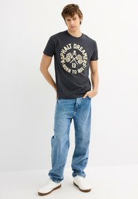 Camiseta de algodón gris oscuro con un estampado gráfico de banderas a cuadros cruzadas y una calavera. Combinada con jeans de mezclilla azul claro y zapatillas blancas.