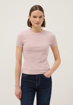 Vrouw met kort bruin haar die gouden oorringen draagt, een lichtroze aansluitend T-shirt met korte mouwen en donkerblauwe jeans, met één hand in de zak.