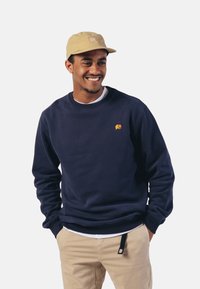 Sudadera de algodón azul marino con un pequeño logo de elefante naranja, combinada con una gorra beige. El modelo lleva pantalones beige con un detalle de bolsillo.