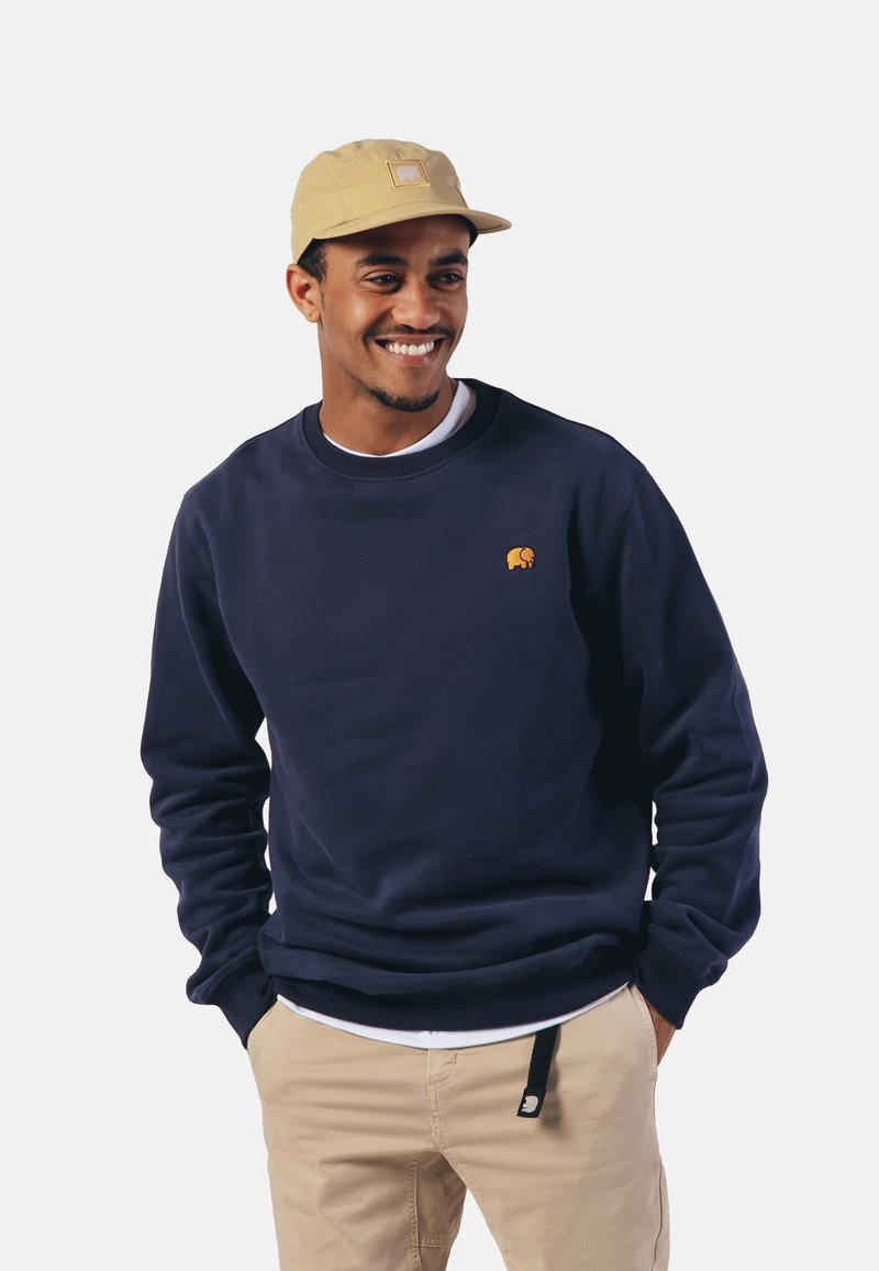 Sudadera de algodón azul marino con un pequeño logo de elefante naranja, combinada con una gorra beige. El modelo lleva pantalones beige con un detalle de bolsillo.