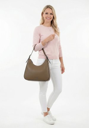 Donna sorridente, indossa un maglione rosa chiaro, pantaloni bianchi, scarpe da ginnastica bianche, con una grande borsa a tracolla in pelle marrone sul braccio sinistro.