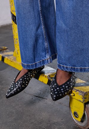 Person iført blå jeans med vide ben og sorte spidse flats med metalnitter, fødderne hviler på en gul og sort industriel barriere.