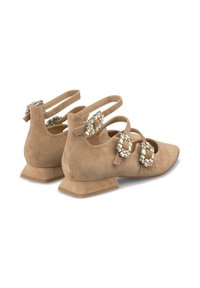 Zapatos planos de ante beige con adornos de cristal decorativos, correas ajustables y un tacón cuadrado bajo. Textura suave y diseño de punta redondeada.