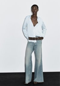 Light blue button-up shirt, dark brown belt with round buckle, faded wide-leg jeans, and black sandals against a plain background.