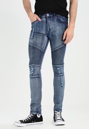 Mann trägt eng anliegende, ausgewaschene blaue Jeans mit gerippten Kniepolstern und schwarze High-Top-Sneaker, die Hände in den Taschen ruhend.