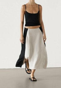 Zwarte mouwloze top gecombineerd met een beige rok tot op de knie met zwarte zijpanelen. Sandalen maken de outfit compleet en tonen een minimalistische stijl.