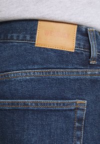 Jeans de mezclilla azul oscuro con una etiqueta de cuero marcada "WEEKDAY", costuras naranjas y diseño clásico de cinco bolsillos.