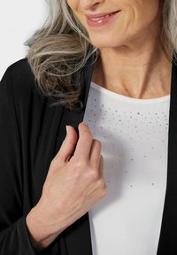 Schwarze Strickjacke über einem weißen Oberteil, das mit kleinen, glitzernden Strasssteinen verziert ist und eine glatte Textur sowie ein klassisches Design zeigt.