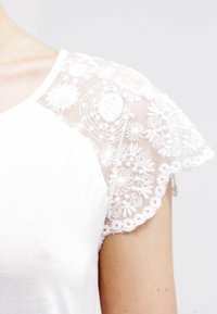 Biały bawełniany t-shirt z krótkimi, przezroczystymi koronkowymi rękawami z haftem kwiatowym i falbaniastymi brzegami, tworzącymi teksturowany kontrast.