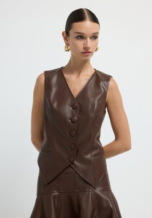 Donna con capelli slicked-back, che indossa un gilet in pelle marrone senza maniche, abbottonato, e una gonna coordinata, accessorista con grandi orecchini a cerchio in oro.