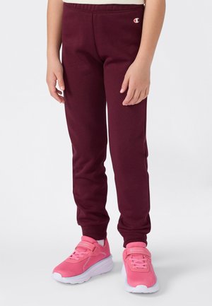 Un enfant portant un pantalon de survêtement bordeaux et des chaussures de sport roses vif se tenant sur un fond blanc.