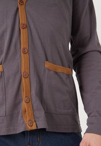 Cárdigan gris de manga larga con ribete de microterciopelo marrón en el cierre de botones y en los bolsillos; presenta botones redondos marrones y una textura suave.