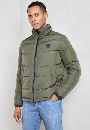Olive green puffer jacket with high collar, quilted design, front zipper, and black logo patch; paired with blue jeans.