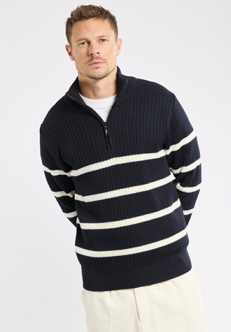 Homme avec les cheveux courts portant un pull côtelé bleu marine avec des rayures horizontales blanches et un pantalon beige, se tenant devant un fond uni.