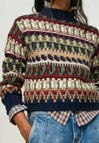 Pull tricoté avec un motif multicolore en vert, rouge, marine et beige. Il présente un col et des poignets côtelés, superposé à une chemise à carreaux.