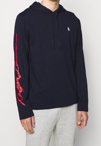 Námořnická košile s dlouhým rukávem a kapucí vyrobená z bavlny, se bílým logem na hrudi a červeným textem v podpisovém stylu podél levého rukávu.