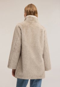 Beige nep bontjas met een hoge kraag, losse pasvorm en zachte textuur. Heeft verlaagde schouders en een rechte zoom, gedragen over een blauwe spijkerbroek.