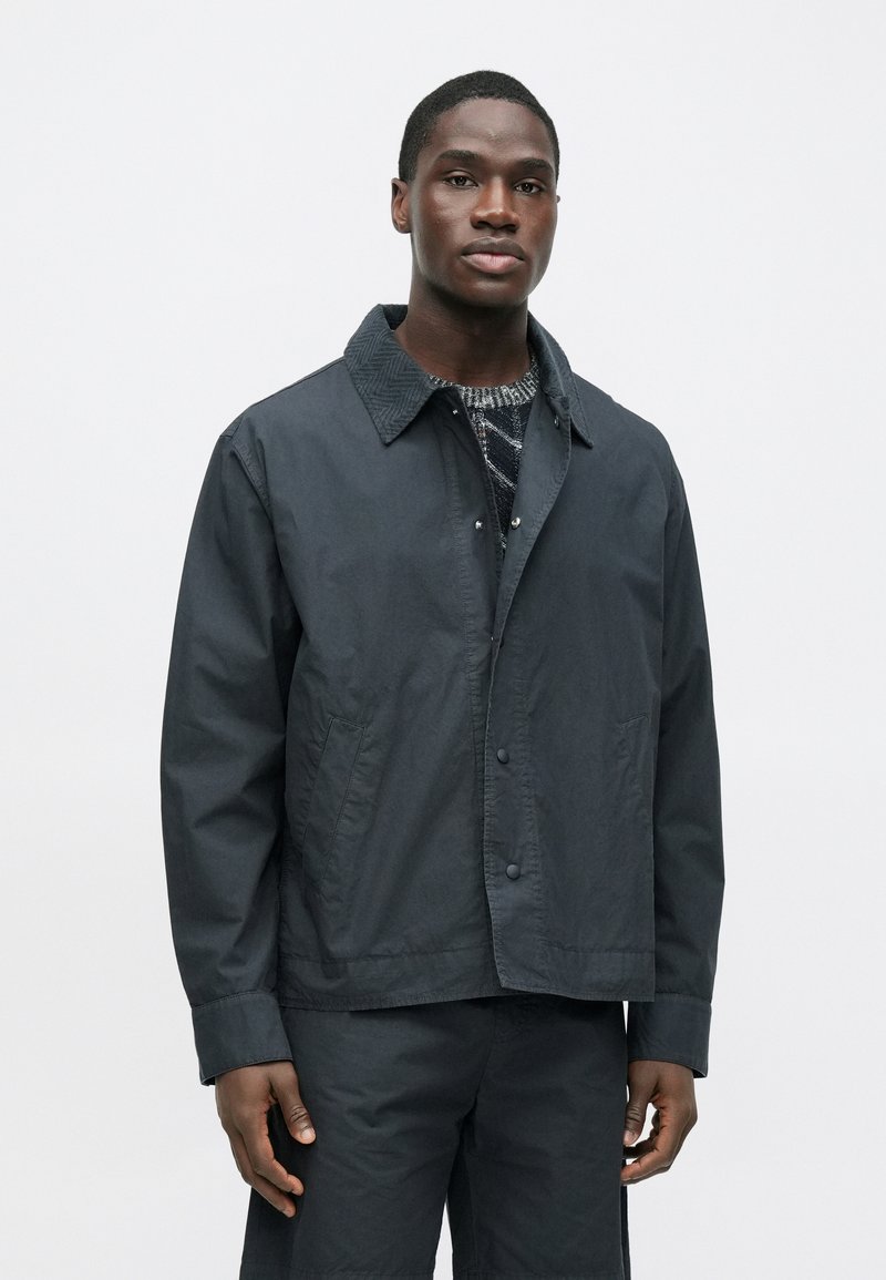 Black lightweight jacket with a textured collar, button closure, and side pockets. Smooth fabric with a minimalist design featured on a model.