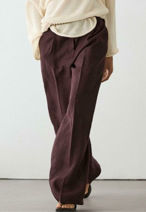 Personne portant un pantalon large ample marron foncé et un pull beige clair, debout pieds nus sur un sol gris devant un fond blanc uni.