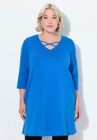Donna bionda che indossa una tunica blu brillante con fasce incrociate al collo a V e maniche a tre quarti, in piedi su uno sfondo bianco.