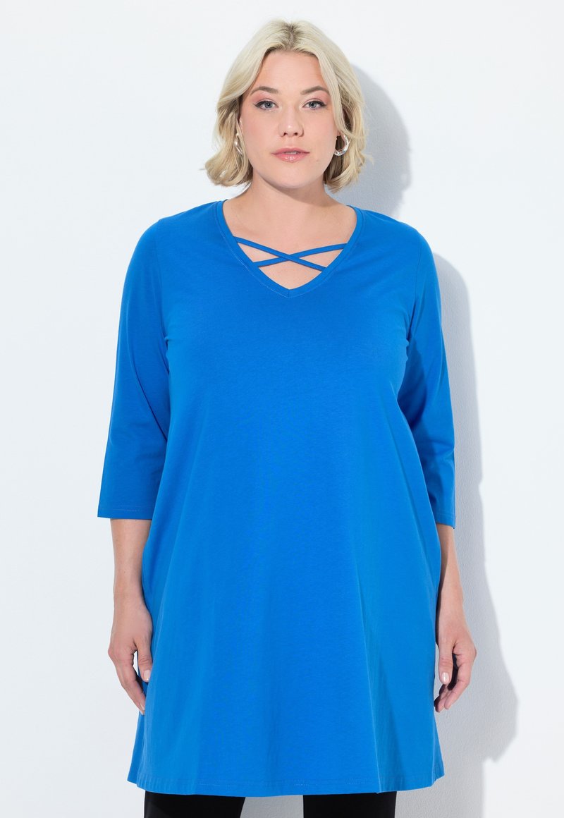 Donna bionda che indossa una tunica blu brillante con fasce incrociate al collo a V e maniche a tre quarti, in piedi su uno sfondo bianco.