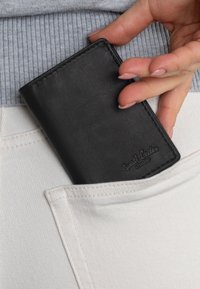 Zwarte leren portemonnee die gedeeltelijk in een witte denimzak is gestoken, met een gladde textuur en een verhoogd logo in de rechteronderhoek.