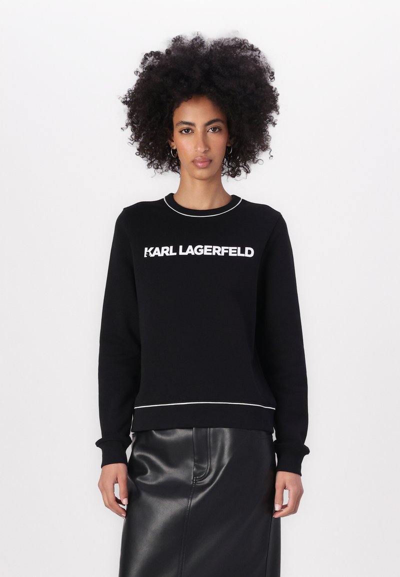 Melnā mikina ar baltu "KARL LAGERFELD" logotipu, ribota apaļa kakla izgriezuma apdare un balts akcenti apakšmalā. Modelis valkā melnu ādas svārku.