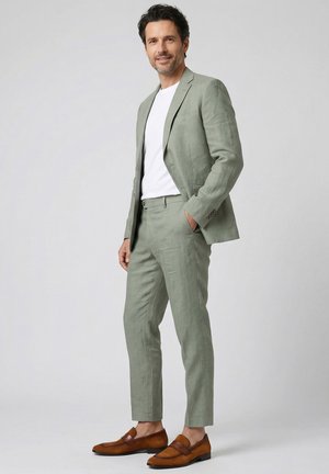 Mann trägt hellgrünen Leinenanzug mit weißem T-Shirt und braunen Ledermokassins, steht mit einer Hand in der Tasche vor schlichtem Hintergrund.