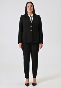 Blazer nero su misura con bottoni argentati, abbinato a pantaloni neri aderenti. Sotto, una blusa crema con un sottile nodo al collo.