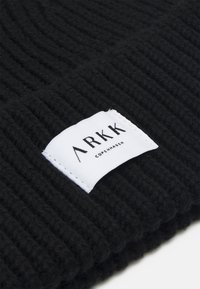 Black knit beanie with ribbed texture. Features a white label with "ARKK COPENHAGEN" printed in black, positioned on the front.
