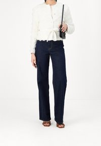 Weißes, strukturiertes, kurzes Jackett mit langen Ärmeln und Bindegürtel. Kombiniert mit dunkelblauen, geraden Jeans und braunen, genoppten Sandalen.
