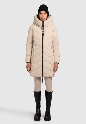 Beige puffer coat met een chevronpatroon, hoge kraag en ritssluiting aan de voorkant. Gecombineerd met zwarte kniehoge laarzen en goed passende lichte broek.