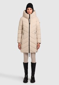 Beige padded coat with chevron quilting, high collar, and front zipper. Worn with black knee-high boots and light leggings. Black knit hat.
