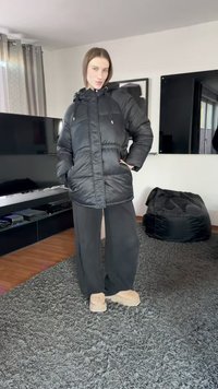 Crna puhovka s kapuljačom, prednjim zatvaračem na patent, dva džepa i prošivenim uzorkom. Kombinirana s širokim crnim hlačama i bež obućom.