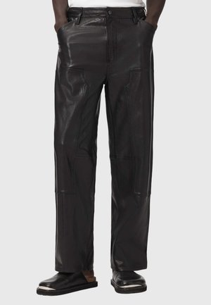 Homme portant un pantalon en cuir noir avec des détails en patch, mains dans les poches, assorti à des chaussures noires, sur un fond clair uni.