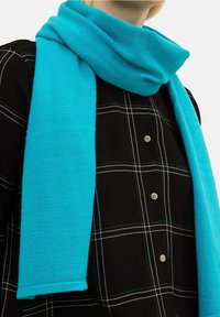 Écharpe turquoise en tissu doux, avec une texture lisse et une forme rectangulaire, drapée autour d'une chemise à carreaux foncés.