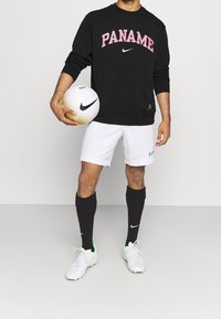 Felpa nera con la scritta "PANAME" in rosa, pantaloni corti bianchi, calze nere e scarpe bianche con dettagli verdi. In mano una palla da calcio bianca e nera.