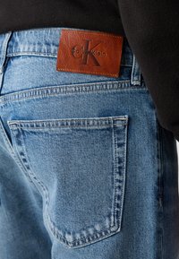 Lyseblå jeans i denim med teksturert finish, som har en brun lærlapp merket "Calvin Klein" på baksiden av linningen.