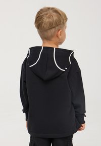 Bambino con capelli biondi corti che indossa un hoodie nero con bordi bianchi sul cappuccio, girato di spalle su uno sfondo neutro.
