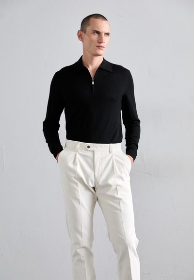 Homem a usar uma camisa preta de manga longa com fecho e uma calça plissada creme de cintura alta, em pé com as mãos nos bolsos contra uma parede lisa.