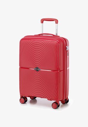 Czerwony twardy bagaż z falistą fakturą, z zamkiem błyskawicznym, przedłużanym uchwytem oraz czterema kółkami dla łatwości poruszania się. Logo marki wyeksponowane w widocznym miejscu.
