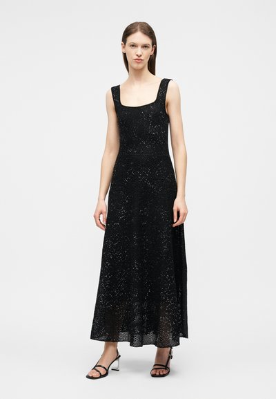 Γυναίκα που φοράει μακρύ μαύρο φόρεμα με παγιέτες χωρίς μανίκια και μαύρα ψηλοτάκουνα σανδάλια με λουράκια, στέκεται μπροστά σε απλό ανοιχτό φόντο.