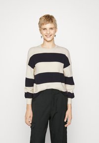 Pull en tricot à rayures crème et marine avec une coupe décontractée, un col rond, des poignets côtelés et une texture douce. Associé à un pantalon noir.