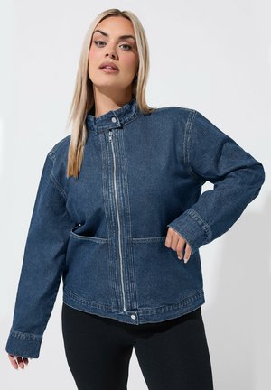 Kvinde med langt blondt hår iført en blå denim lynlåstrøje og sorte bukser mod en ensfarvet lys baggrund.