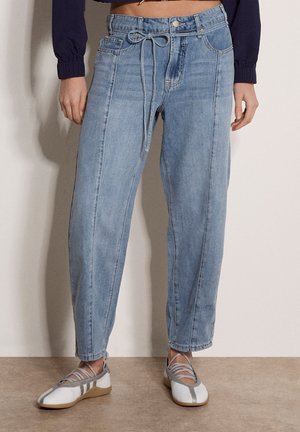 Vrouw die lichtblauwe jeans met hoge taille draagt met een strik aan de voorkant en witte ballerina's met grijze bandjes.